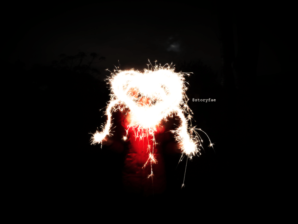 black background person in red coat making heart out of light with sparklers 