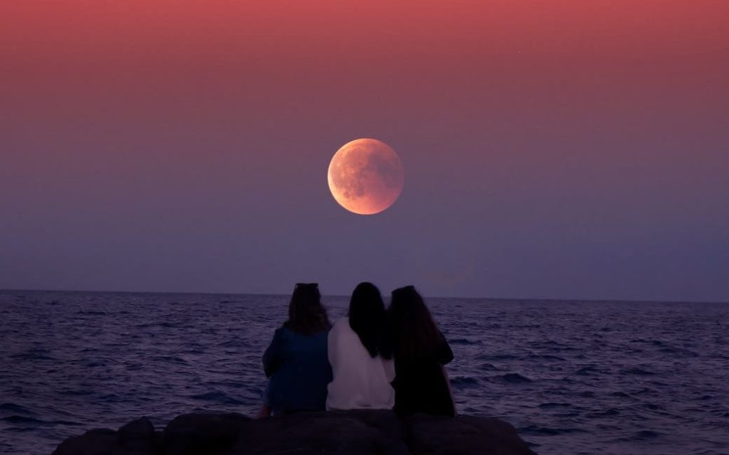 Three women sitting on a rock at the sea shore watching the full moon as it is lid with dramatic sunset colours. 