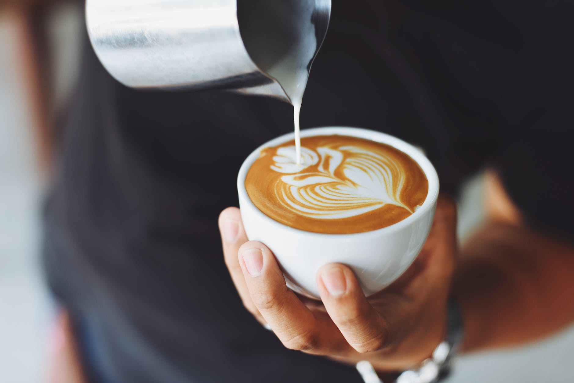 close up of milk foam being artfully poured into a cappuccino. One can see the hand holding the cup, and the turso glad in black pullover in the background, the silver milk jar seems to float at the top of the photo. It's aesthetically pleasing. So much so I want a coffee now. 