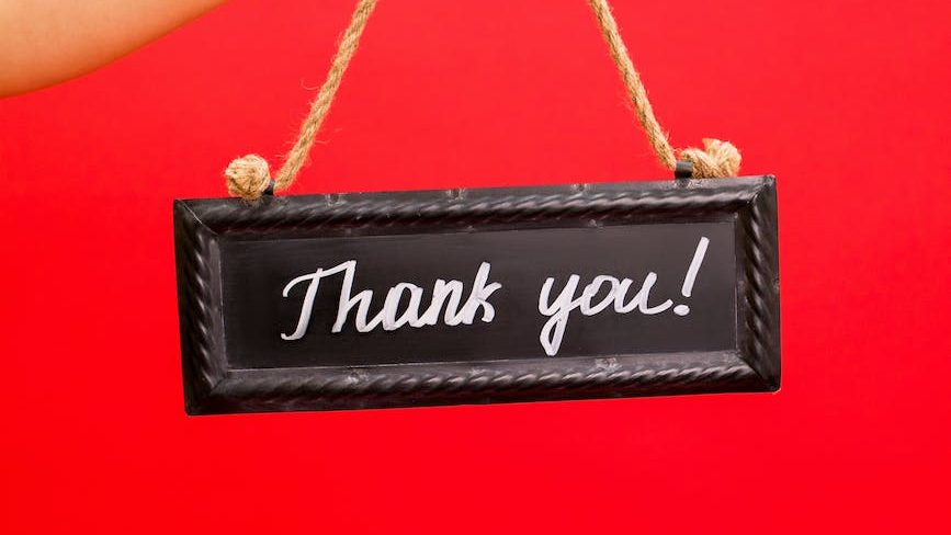 a black sign hanging of rough string, held by a disembodied hand in front of red background. The sign simply says Thank you! (Written in white paint)