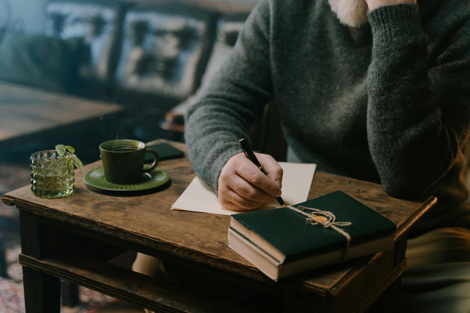 person sitting at a small old wooden table, some journals are balancing at the top right corner edge, the are writing with a fountain pen on a piece of paper, and the left side of the table holds a green ceramic cup and a 70s style green water glass with some mint in it. The person wears a dark green wool pullover and it appears as if they are in a café 